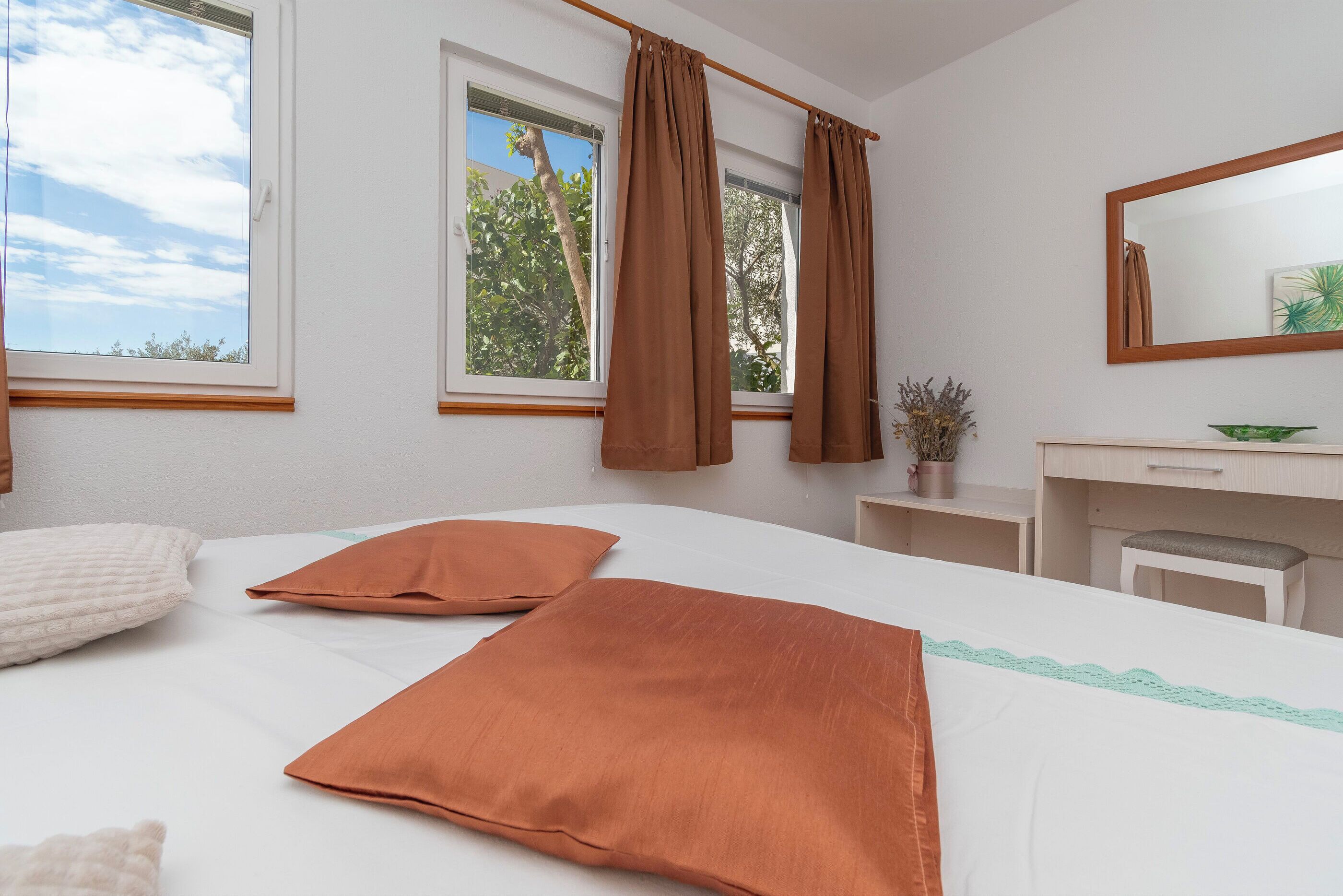 A bedroom with a bed, two pillows, a desk, a chair, a mirror, and a window with curtains.
