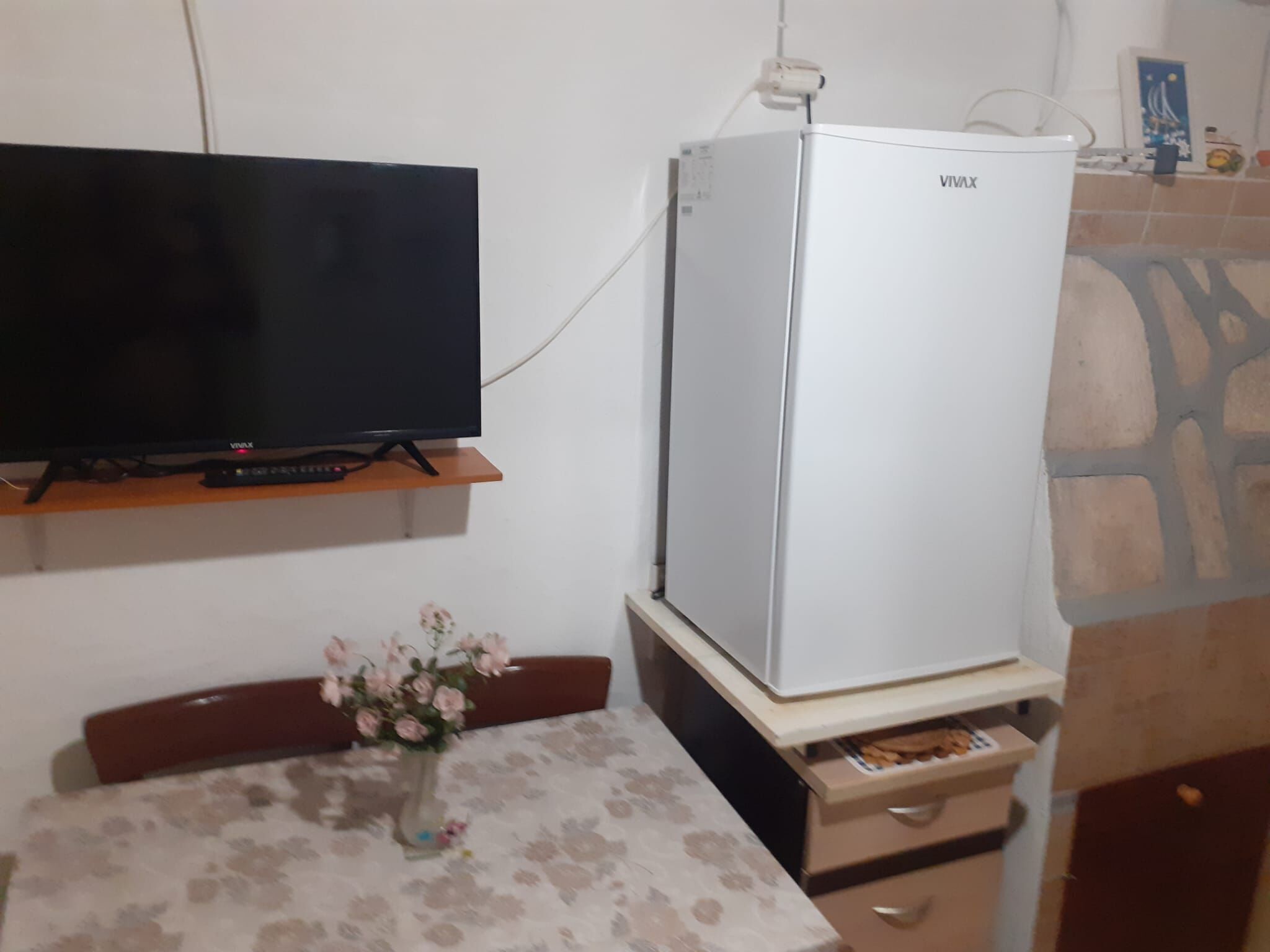 A room with a flat-screen TV on a wooden shelf, a white refrigerator, a floral tablecloth, and a vase with flowers.