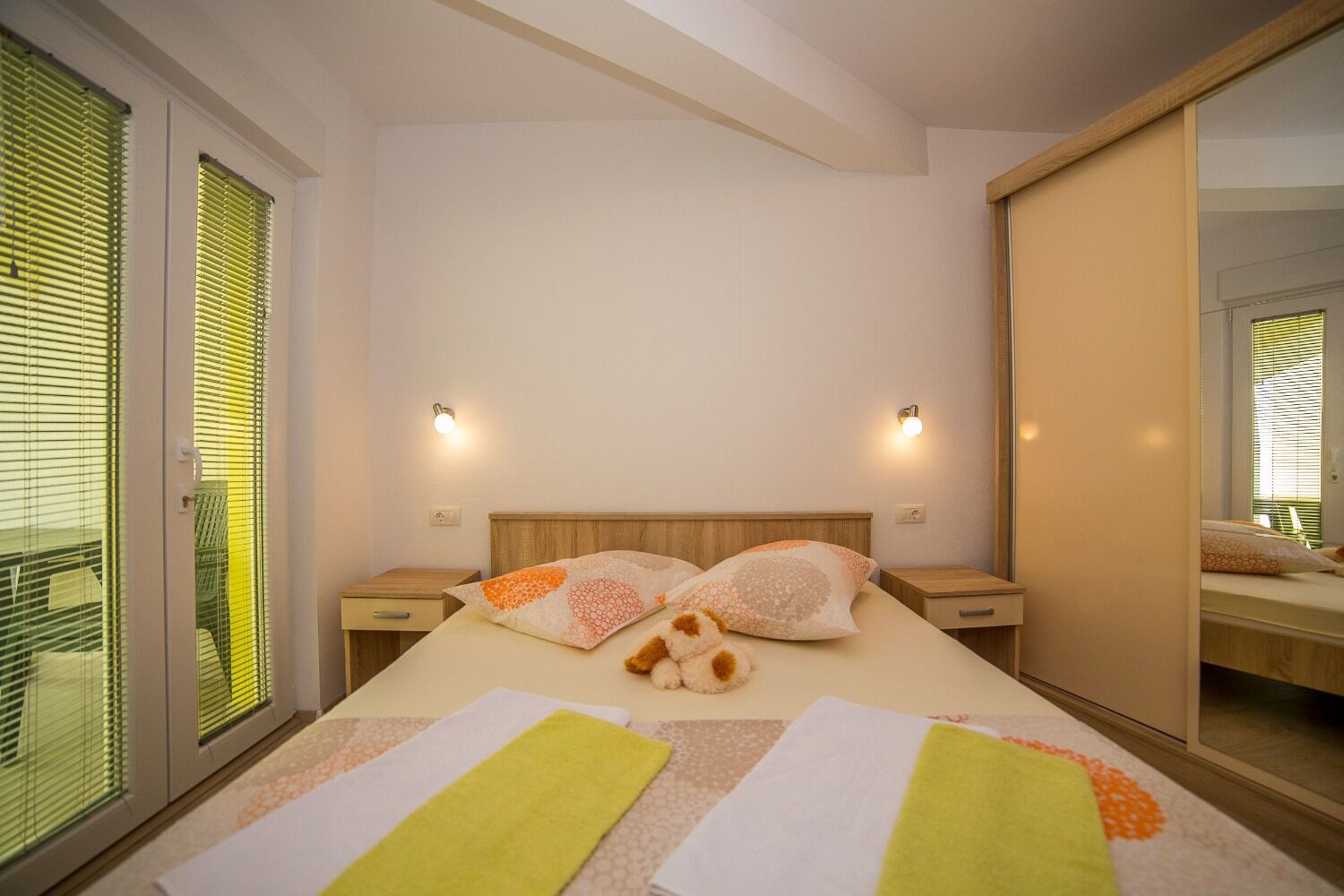 A bedroom with a bed, two bedside tables, a mirror, and a window with blinds.
