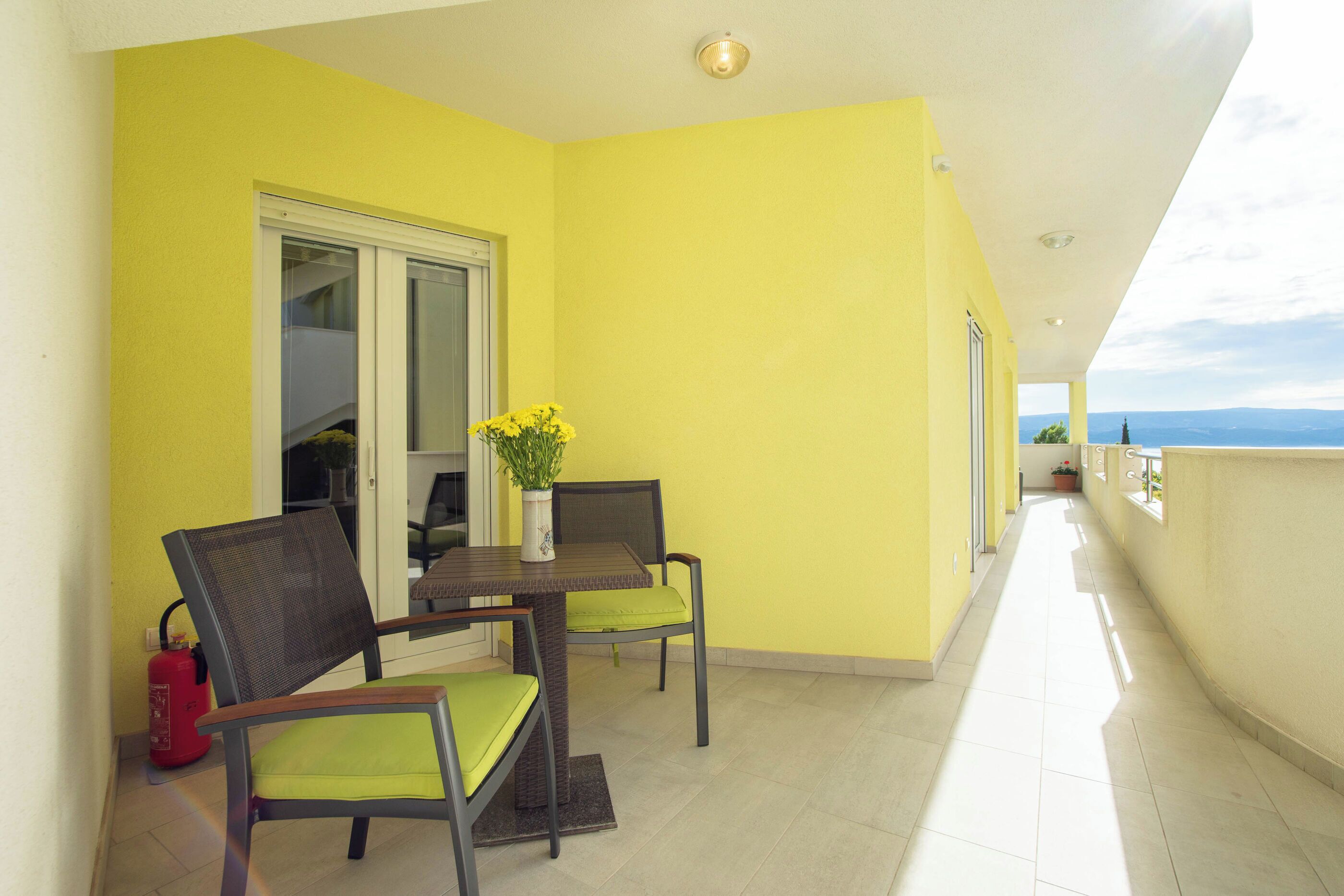 A balcony with yellow walls, a table with chairs, a fire extinguisher, and a view of the landscape.