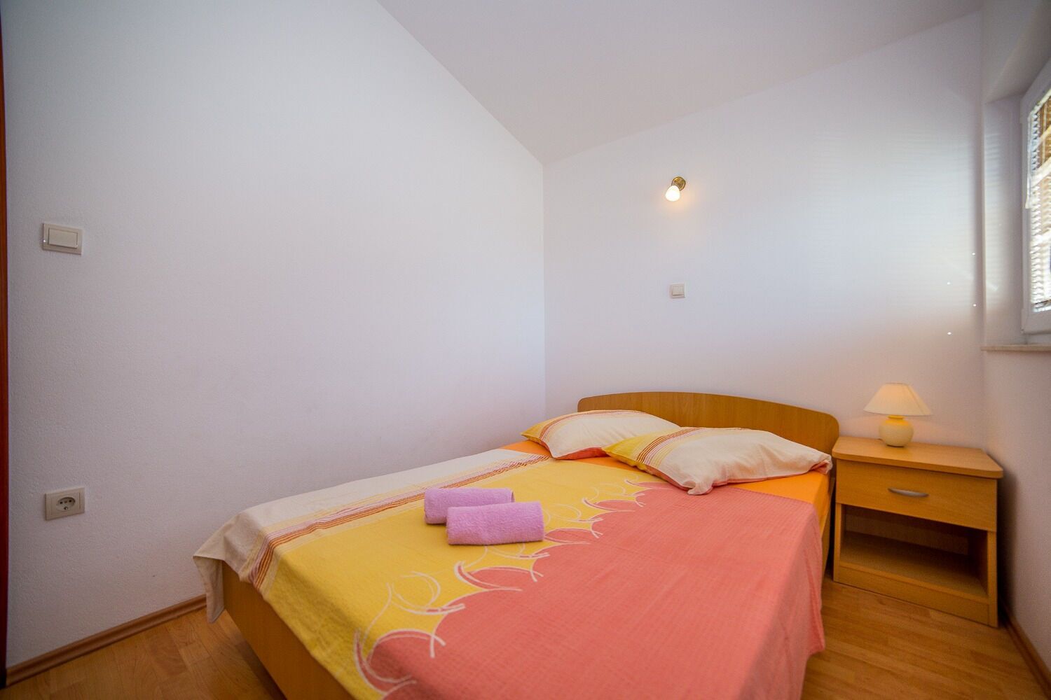 A small, neatly arranged bedroom with a single bed, a bedside table with a lamp, and a power outlet on the wall.