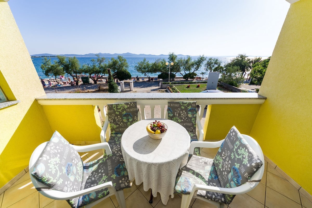 Un balcon avec une table et deux chaises, donnant sur une plage où l’on aperçoit des gens et des arbres.