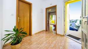Un couloir avec une porte en bois, une plante en pot et une vue sur un balcon où l’on aperçoit une voiture et de la verdure.