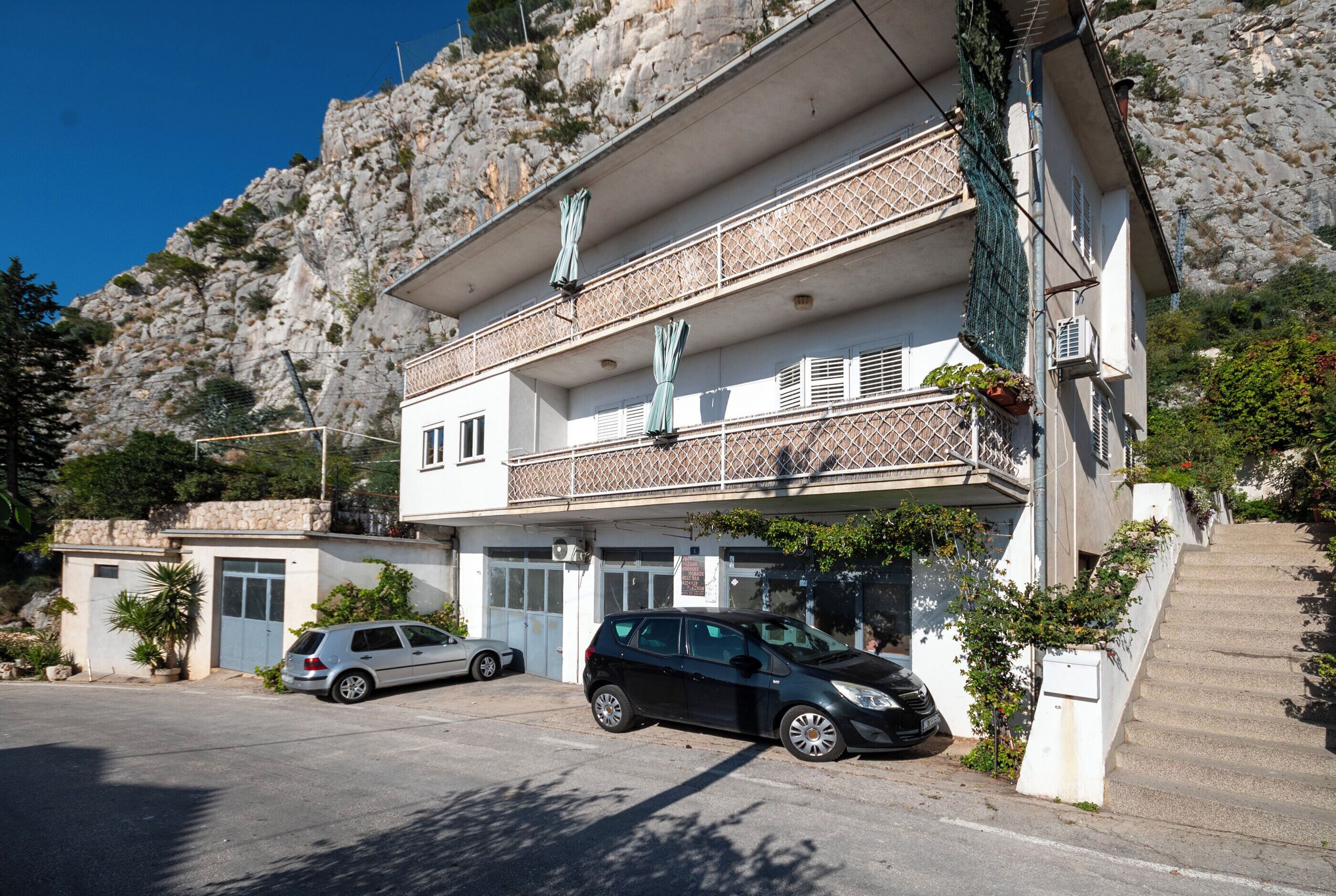 A two-story building with a balcony, a parking area, and a staircase leading to the entrance.