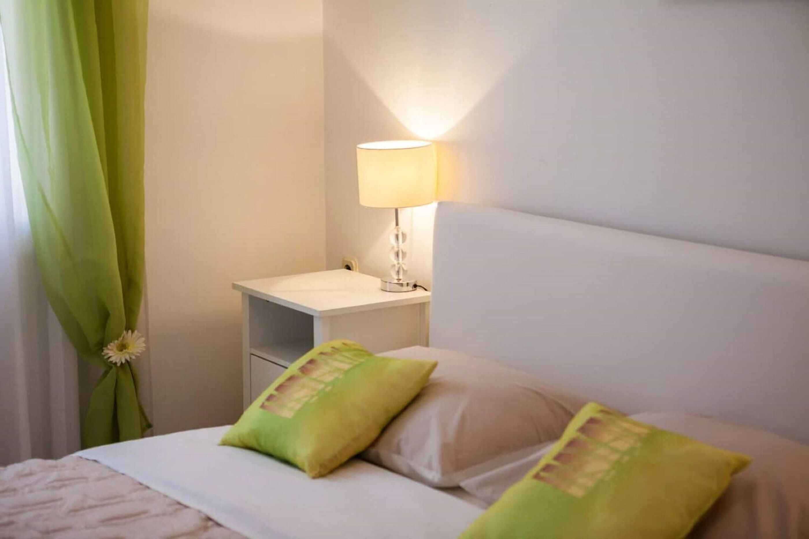Una cama bien tendida con dos almohadas, una mesita de noche con lámpara y una cortina verde con un estampado floral.