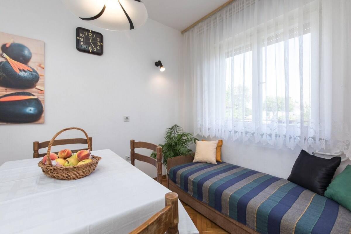 Ein gemütliches Zimmer mit Esstisch, einem Bett mit gestreifter Bettwäsche, einer Wanduhr und einem Fenster mit durchsichtigen Vorhängen.