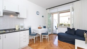 Een kleine keuken met witte kasten, een fornuis en een koffiezetapparaat. Er is een eethoek met een tafel en stoelen, en een donkerblauwe bank in de woonkamer.