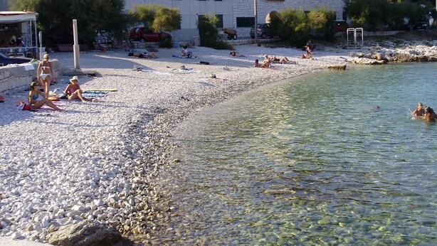 Een kiezelstrand met helder water, mensen aan het zonnen en een gebouw op de achtergrond.