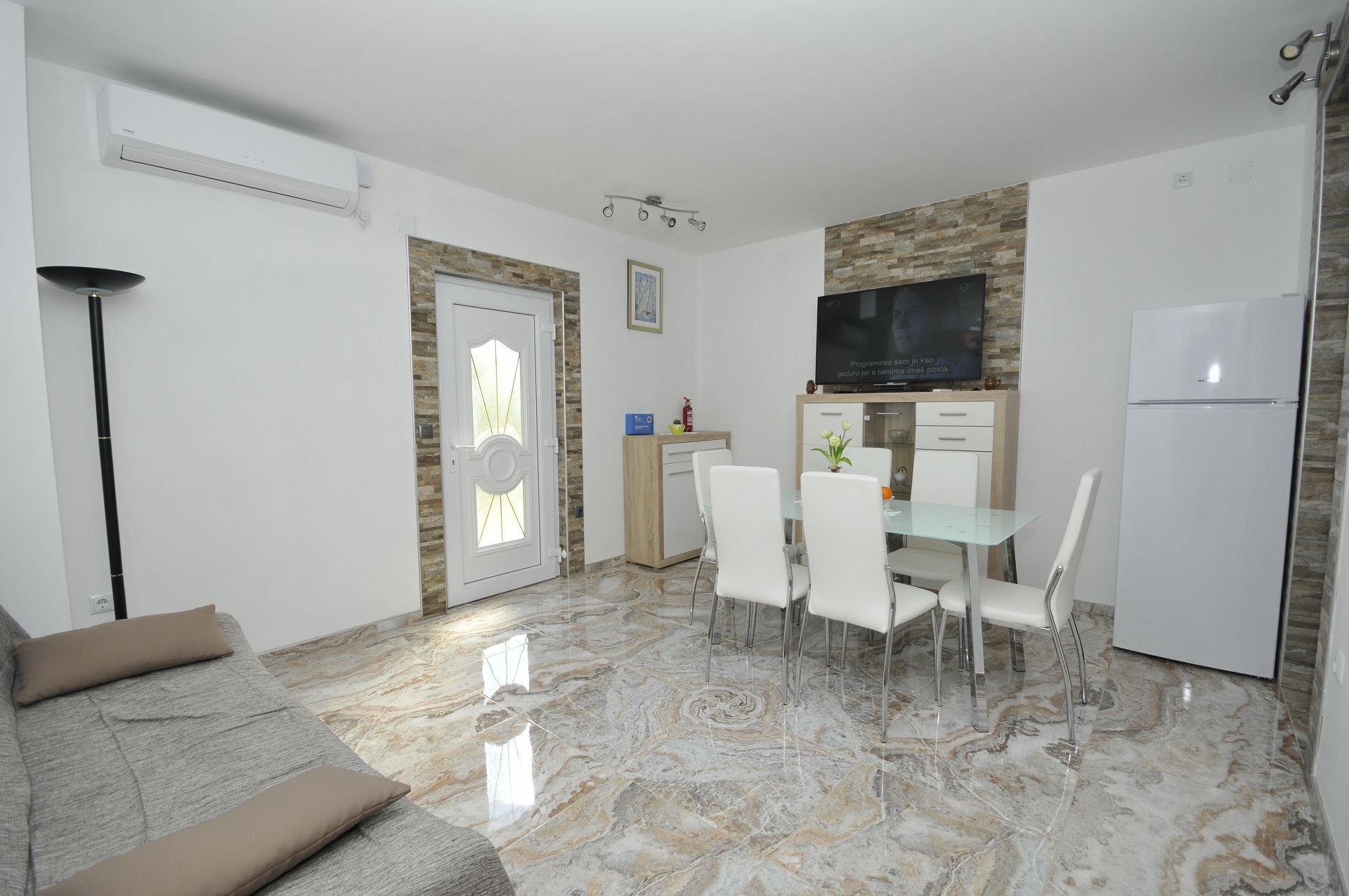 A modern living room with a sofa, a dining area with chairs and a table, a kitchen with a refrigerator, and a TV mounted on the wall.
