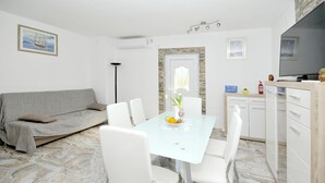 A modern living room with a grey sofa, white chairs, a glass dining table with a fruit bowl, and a kitchen area with white cabinets and a stove. - Apartments Ivica (Kastela)