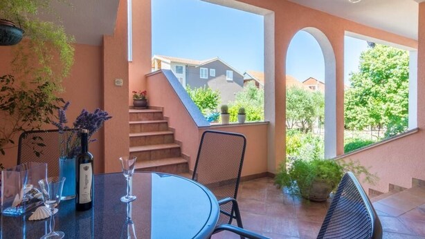 A patio area with a table, chairs, and a bottle of wine. There are stairs leading to a balcony with a view of houses and trees. - Apartment Marinka (Zadar)