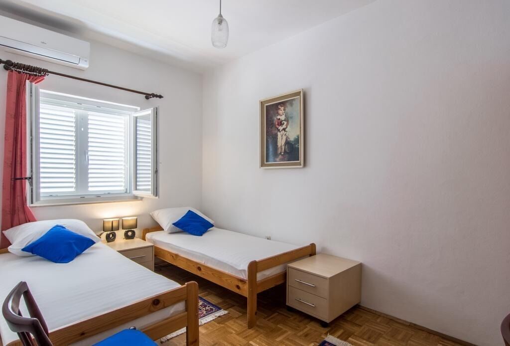 A small, clean room with two single beds, a desk, a chair, and a wall-mounted air conditioner.