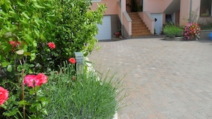 A courtyard with a brick pathway, a building with a staircase, and various plants and flowers. - Apartment Marinka (Zadar)