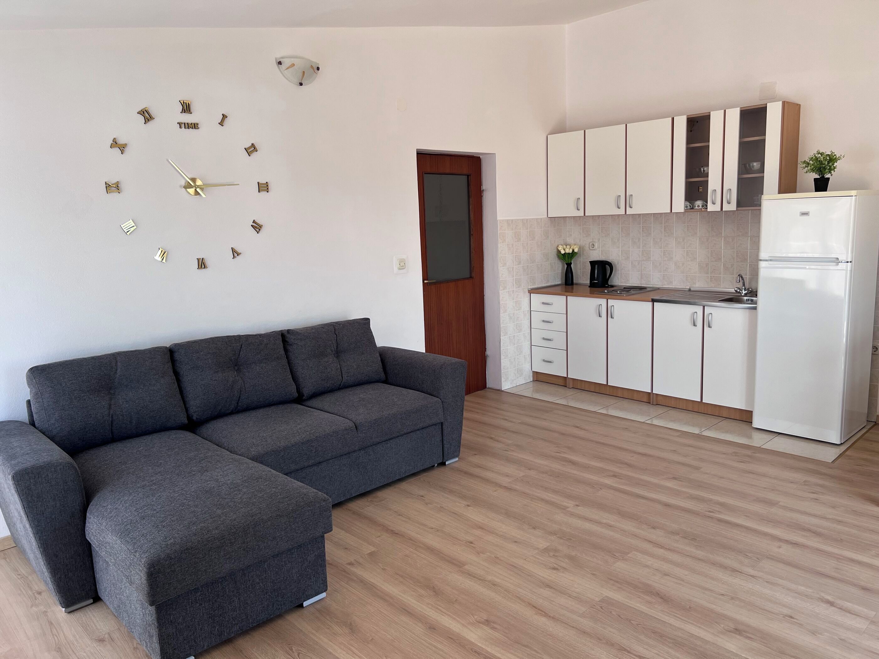 Ein modernes Wohnzimmer mit grauem Sofa, eine Küche mit weißen Schränken und einer Wanduhr.