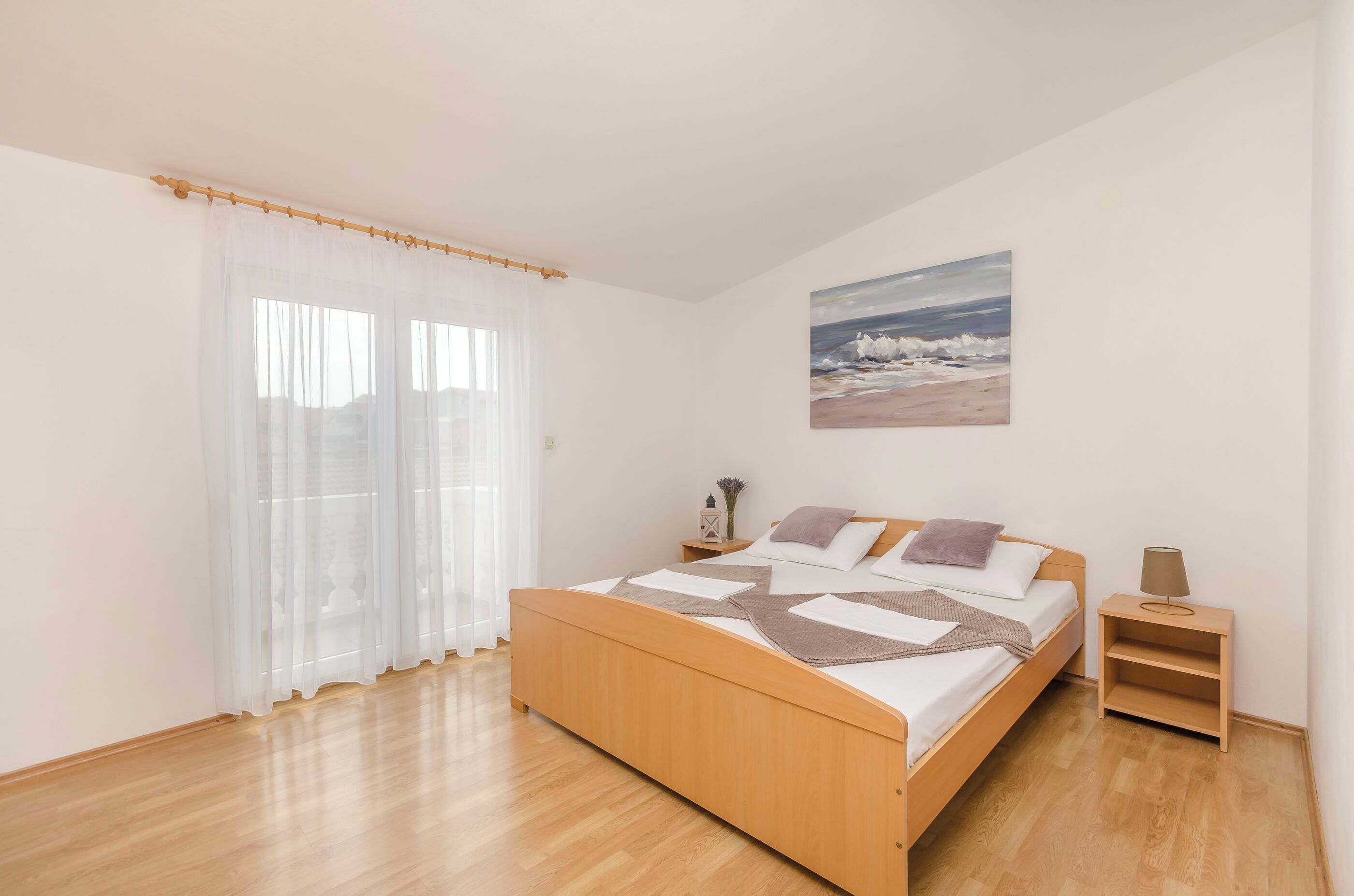 Un dormitorio con cama de madera, cortinas blancas, una mesita de noche, una lámpara y un cuadro con una playa.