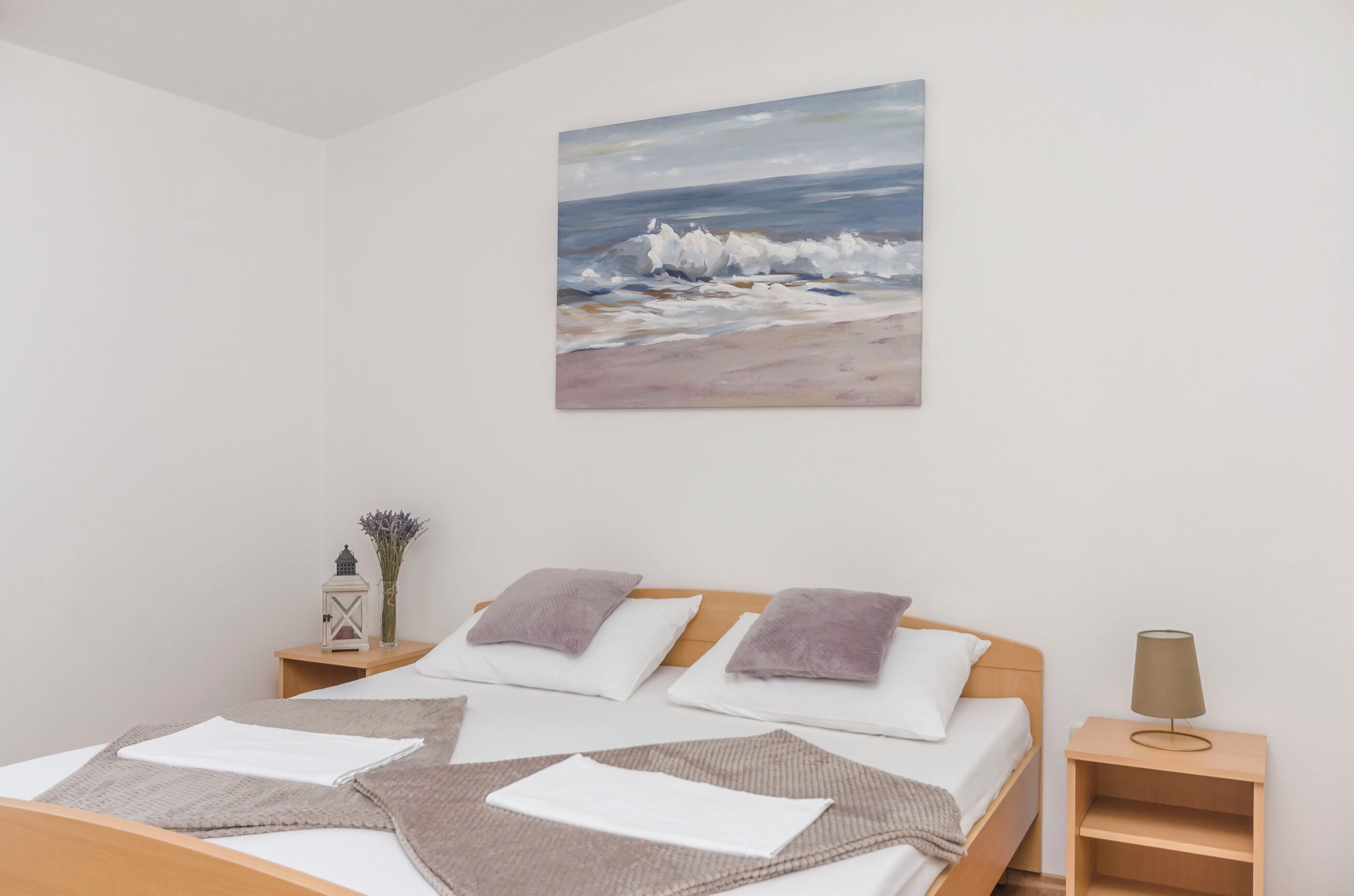A bedroom with a bed, pillows, a nightstand, a lamp, a painting of a beach scene, and a vase with flowers.
