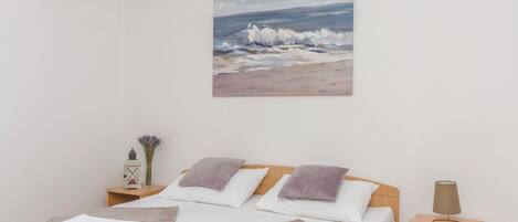 A bedroom with a bed, pillows, a nightstand, a lamp, a painting of a beach scene, and a vase with flowers.