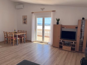Ein Wohnzimmer mit einem Holz-TV-Schrank, einem Esstisch mit Stühlen und einer Glastür zu einem Balkon.