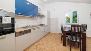 A modern kitchen with a dishwasher, oven, and blue cabinets. There is a dining table with chairs and a window with a view of greenery. - Apartment Toma (Karlobag)
