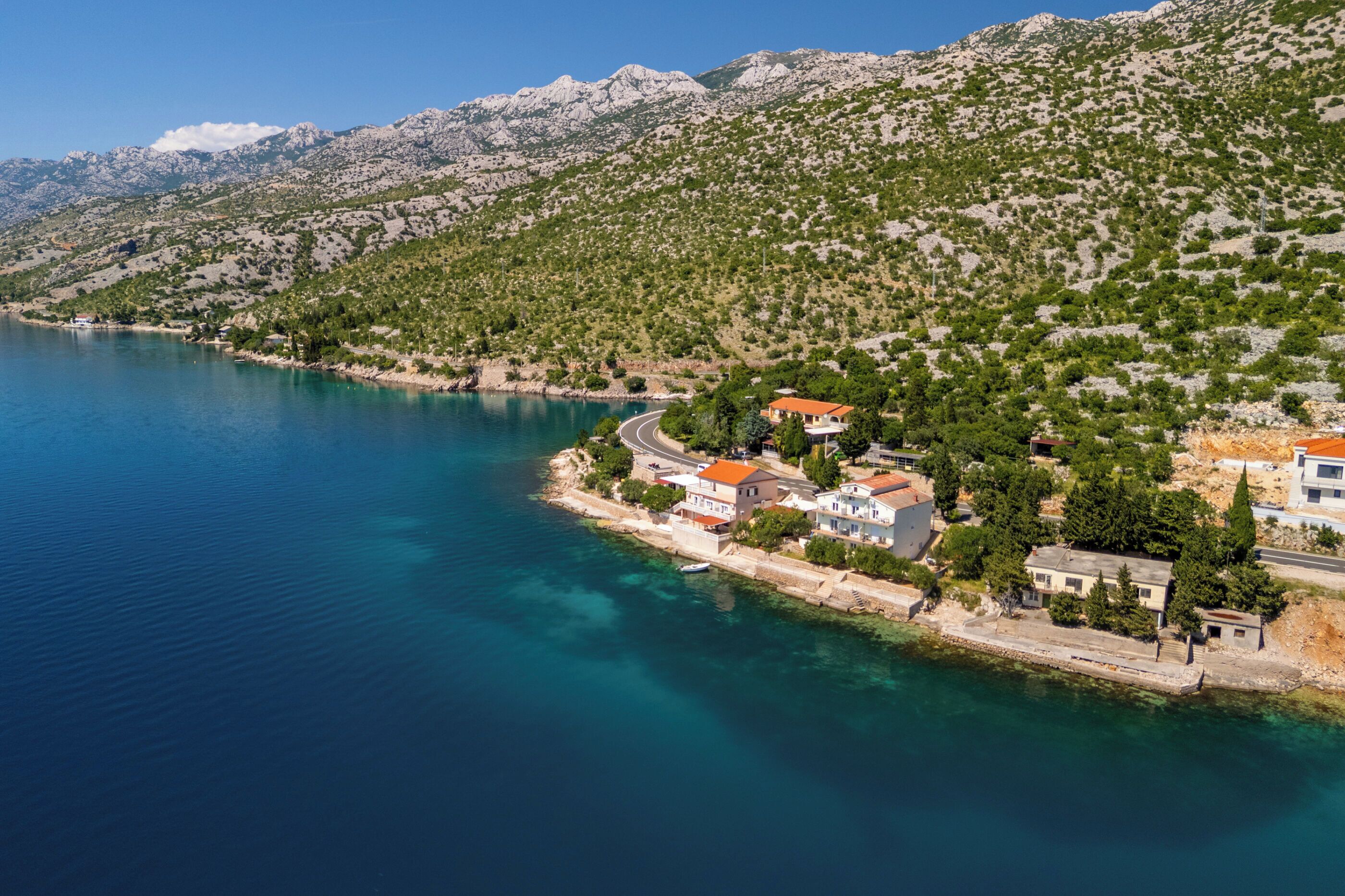 A coastal village with houses along the shoreline, a clear blue sea, and mountains in the background.