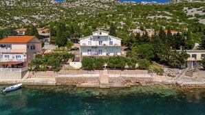 A coastal property with a stone staircase leading to a white building with balconies, situated by clear blue waters and surrounded by greenery and hills. - Apartment Toma (Karlobag)