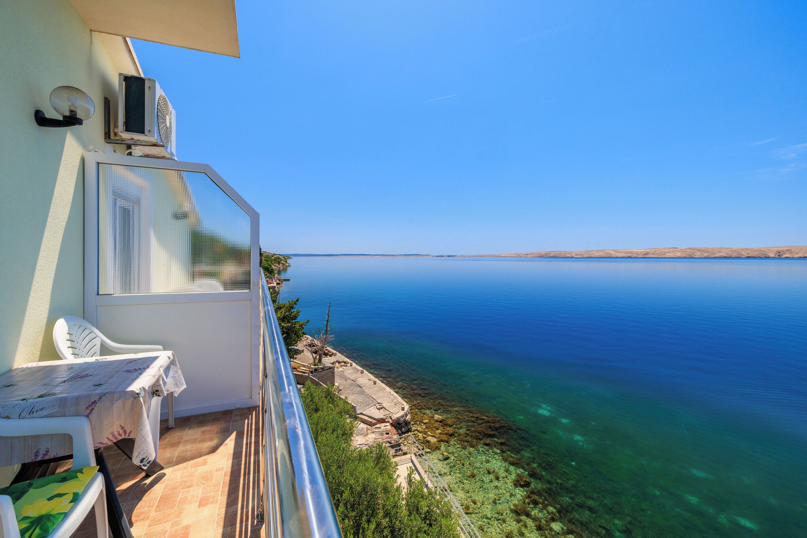 A balcony with a table and chairs, overlooking a clear blue sea and rocky shoreline.