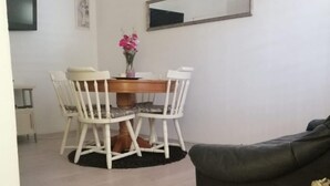 Zona pranzo con tavolo in legno e sedie bianche, divano nero e un vaso con fiori secchi.