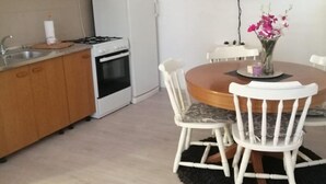 Una cucina compatta con piano cottura, frigorifero e mobili bianchi. Un tavolo da pranzo con sedie e un vaso di fiori si trova al centro.