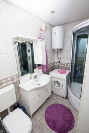 Egy kis méretű fürdőszoba, amelyben mosdókagyló, tükör, WC, mosógép és zuhanyzó található.