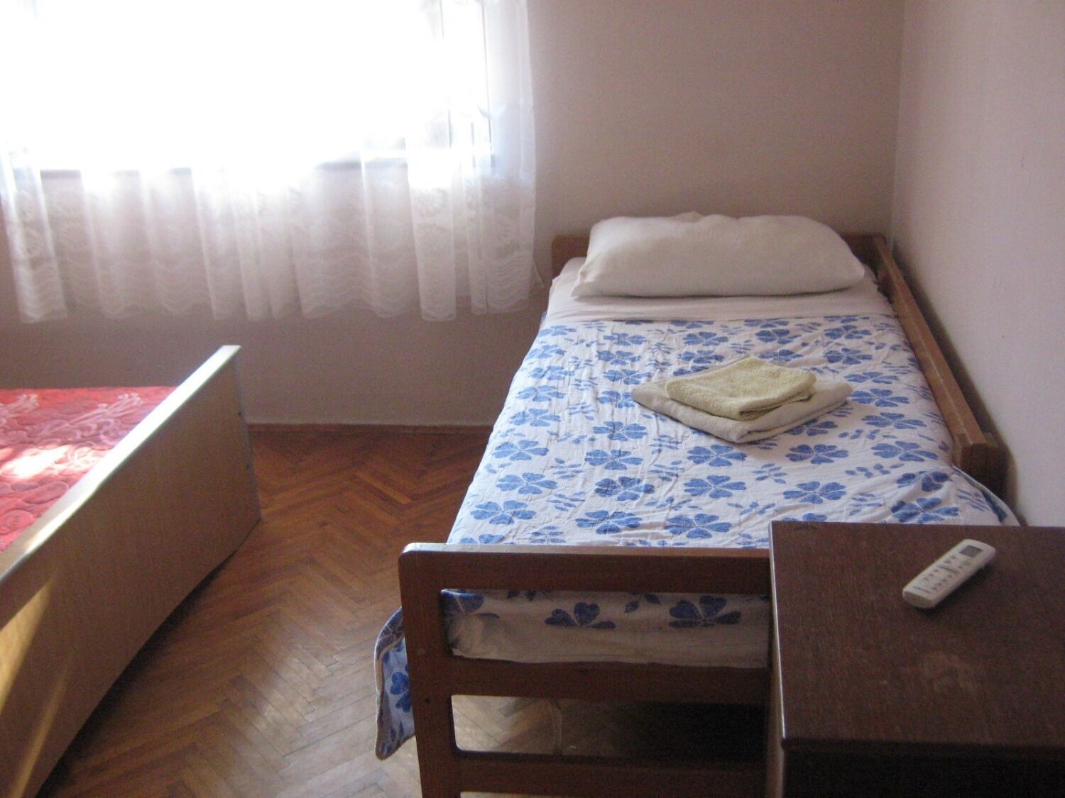Ein kleines Zimmer mit einem Einzelbett, Holzboden, einem Bett mit blumigem, blauem Überwurf, einem weißen Kopfkissen, einem Holzstuhl und einem Nachttisch mit Fernbedienung.