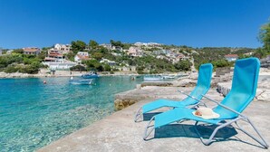 A coastal scene with clear blue water, two blue lounge chairs on a stone platform, and buildings in the background. - Apartment Bosiljka (Marina)