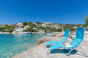 A coastal scene with clear blue water, two blue lounge chairs on a stone platform, and buildings in the background. - Apartment Bosiljka (Marina)