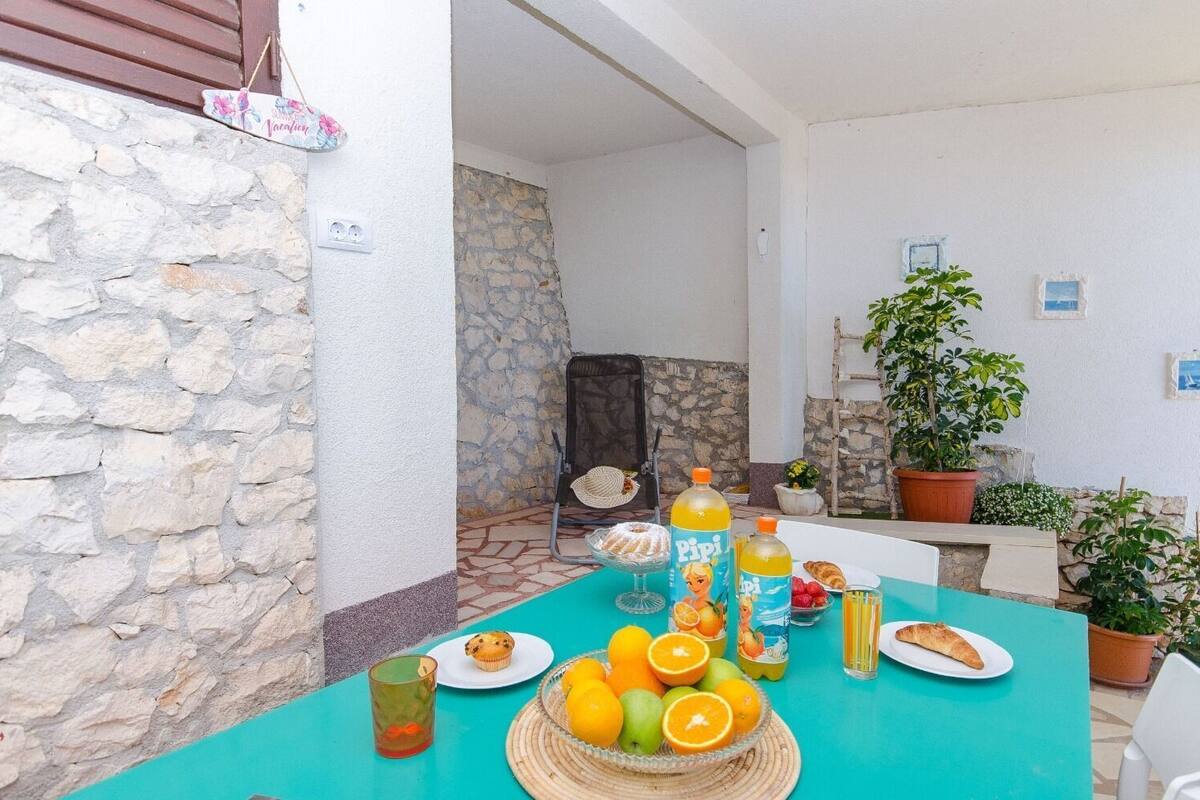 A turquoise table set with fruits, a muffin, and a bottle of Pipi juice, surrounded by stone walls and potted plants.