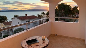 Ein Balkon mit Blick auf einen Sonnenuntergang über dem Wasser, ein Fliesenboden und ein kleiner Tisch mit einem Laptop und einer Kerze.