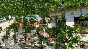An outdoor patio with a round table and white plastic chairs, surrounded by potted plants and a metal fence. - Apartments Josip (Rogoznica)