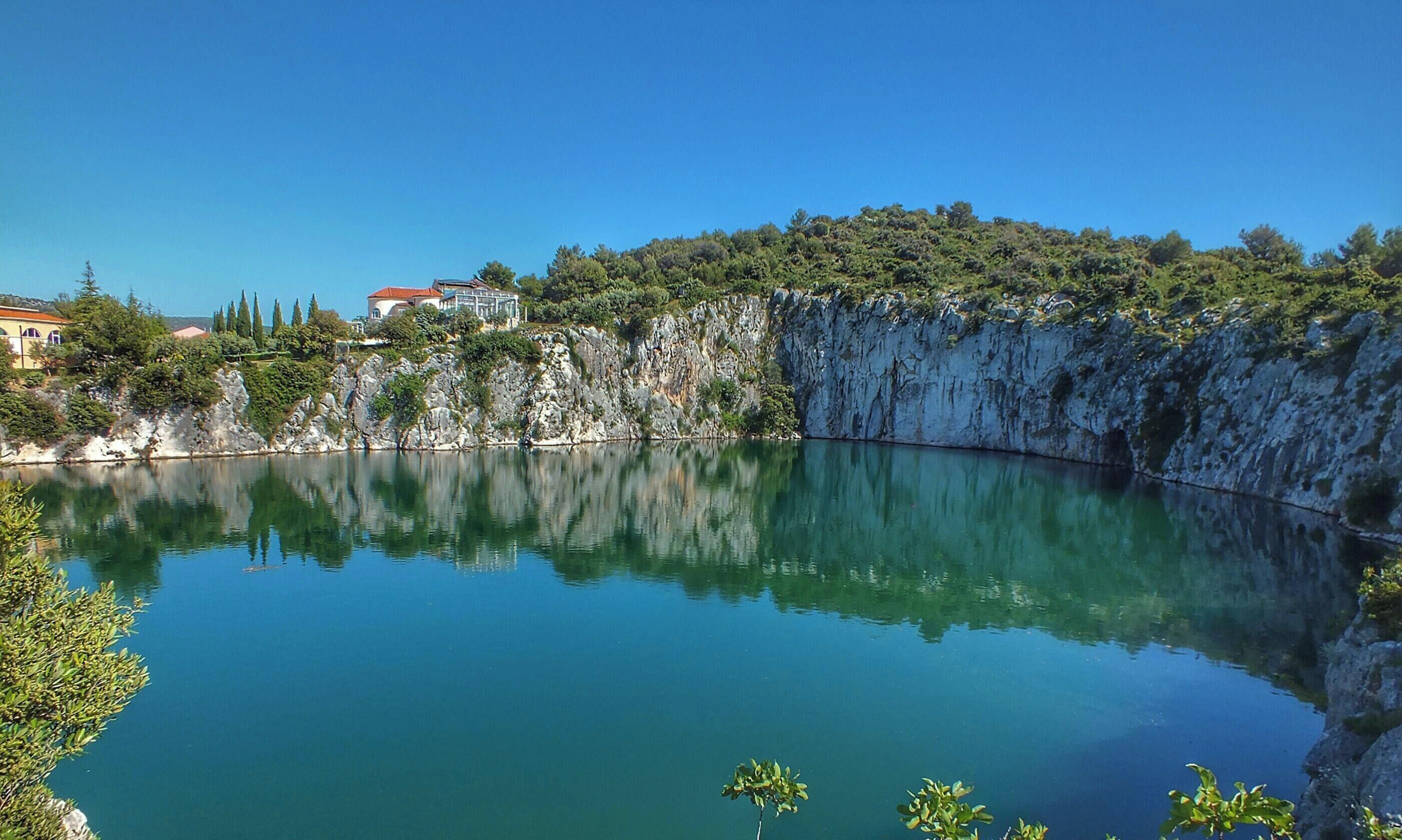 Ein See mit einem Haus auf einer Klippe, umgeben von viel Grün.