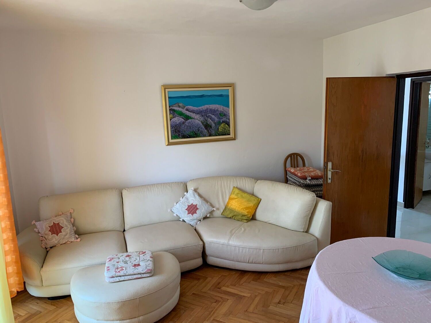 A living room with a beige sectional sofa, a round ottoman, a small table with a green object, and a painting of a landscape on the wall.