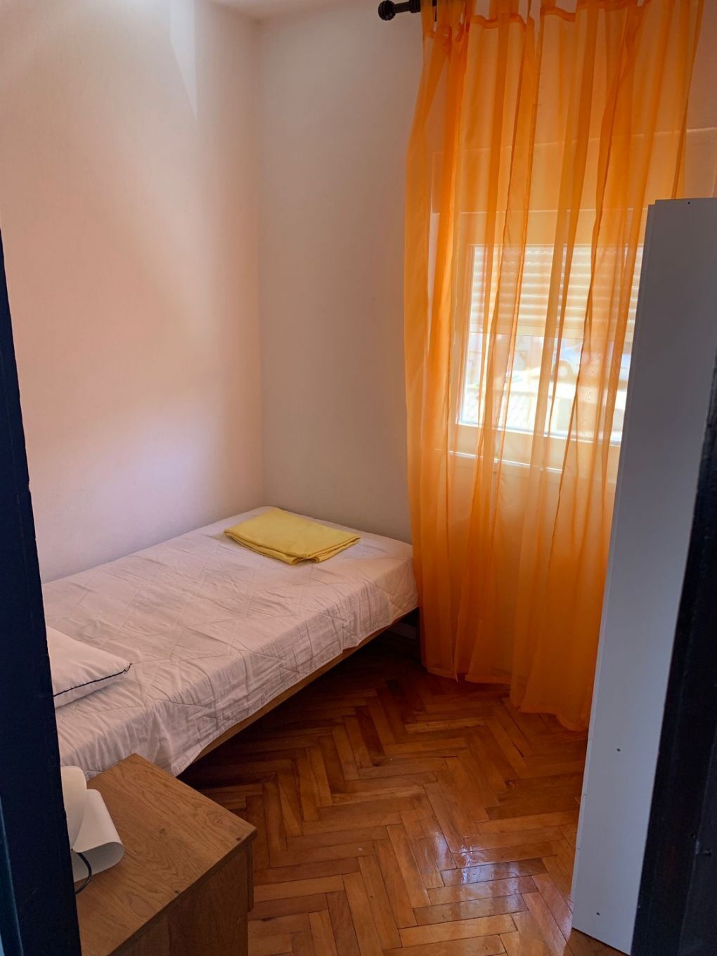 Una camera piccola con un letto singolo, pavimento in legno a spina di pesce, un comodino con una lampada e una finestra con tende arancioni.