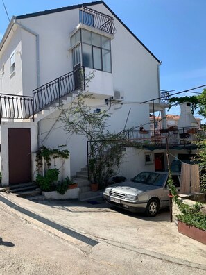 Ein weißes, zweistöckiges Gebäude mit Balkon, einem geparkten Auto davor und einem kleinen Garten.
