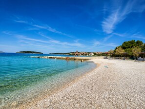 A pebble beach with clear water, a pier, and a distant island. - Apartments Josip (Rogoznica)