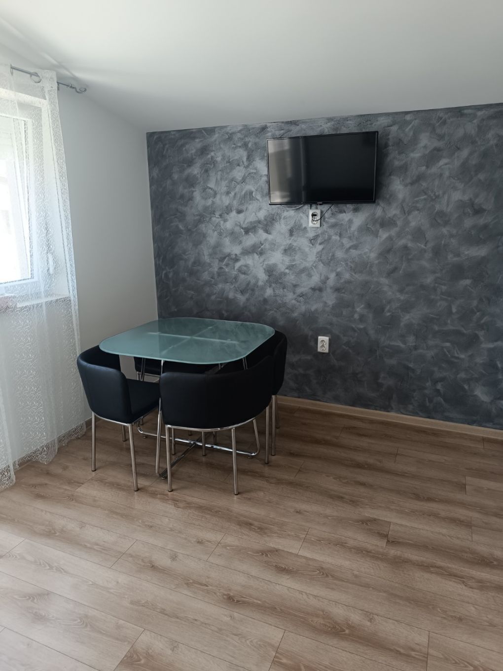 A dining area with a table and chairs, a flat-screen TV mounted on the wall, and a window with a curtain.