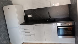 Cocina compacta con armarios blancos, encimera de mármol negro, horno empotrado y refrigerador.