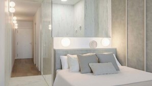 Een slaapkamer met een grote spiegel, een bed met witte lakens, en een gang met een deur.