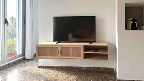 Una sala de estar moderna con un televisor de pantalla plana montado en una consola de madera, un ventanal corredero que da a un balcón y un estante pequeño con objetos decorativos.