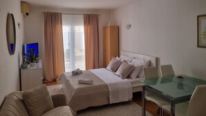 A bedroom with a bed, a desk, and a chair. There is a window with curtains, a mirror, and a wall-mounted light. - Apartment Bubi (Solta)