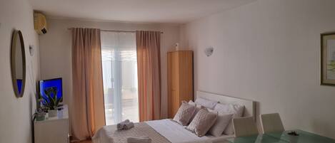 A bedroom with a bed, a desk, and a chair. There is a window with curtains, a mirror, and a wall-mounted light.