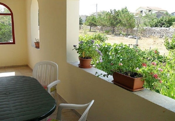 Un balcón con mesa y sillas, plantas en macetas y vistas a un jardín y edificios al fondo.
