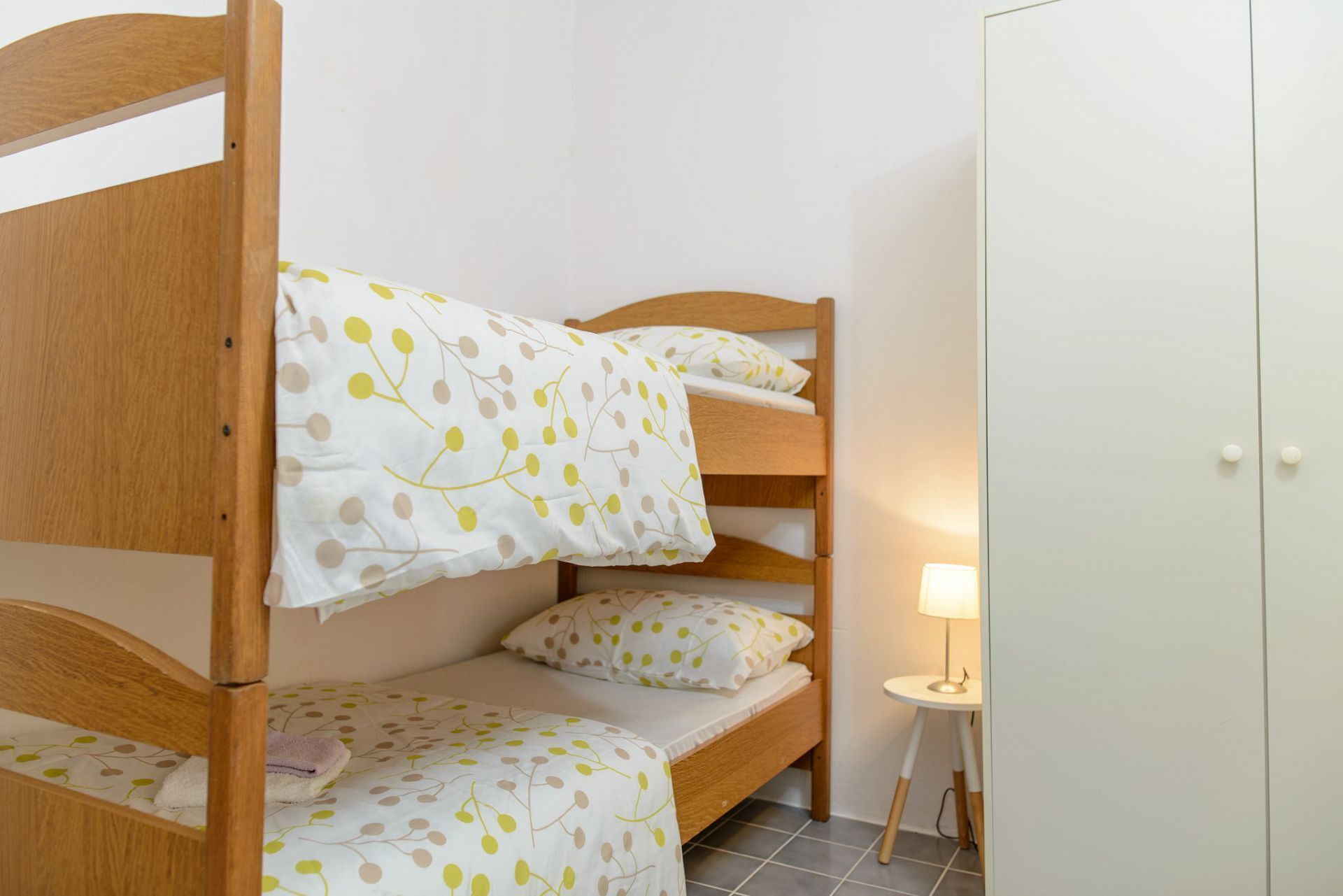A bunk bed with a white and yellow leaf pattern bedspread, a small bedside table with a lamp, and a wardrobe in a room with white walls and grey tiled floor.