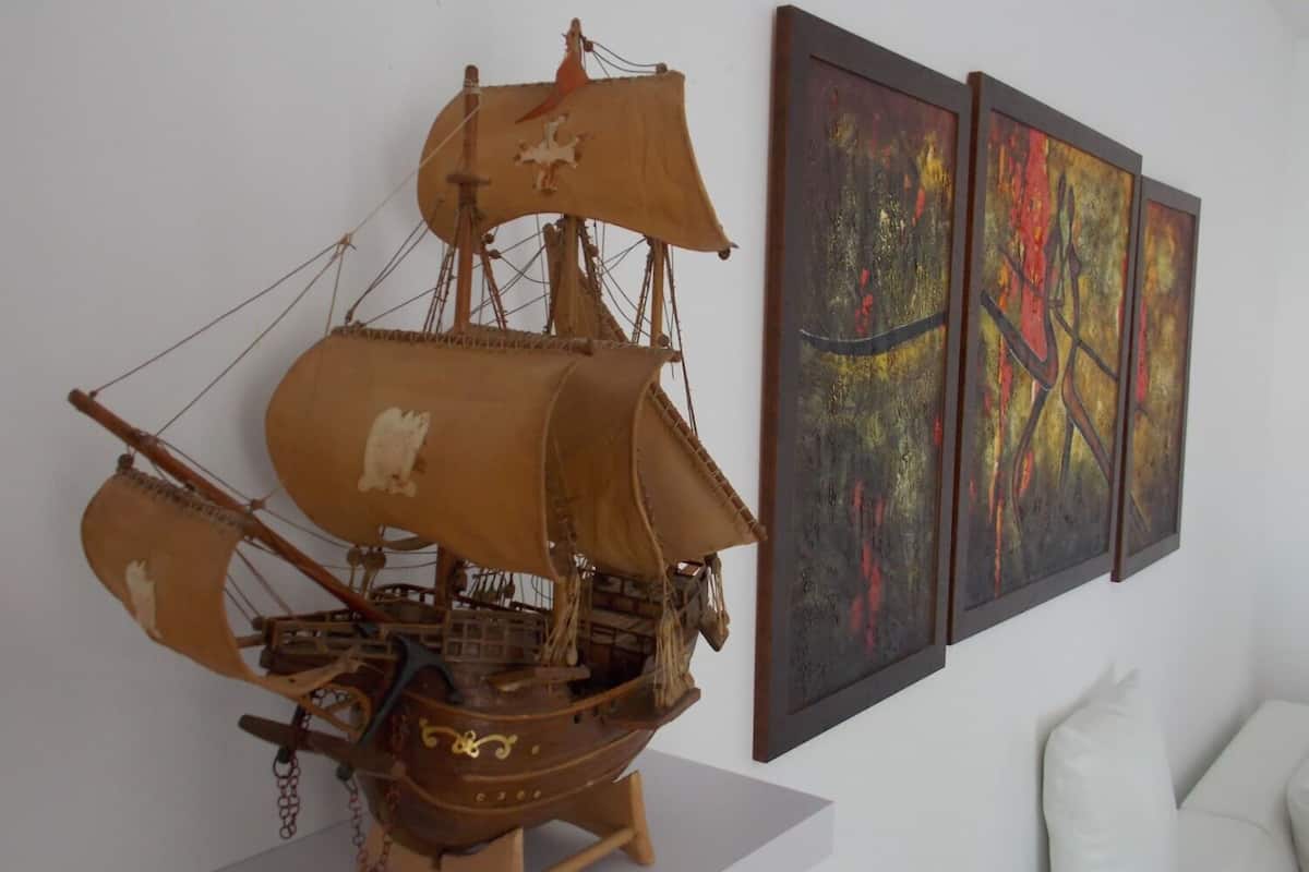 A model ship on display, possibly in a museum or gallery setting, with abstract paintings on the wall to its right.