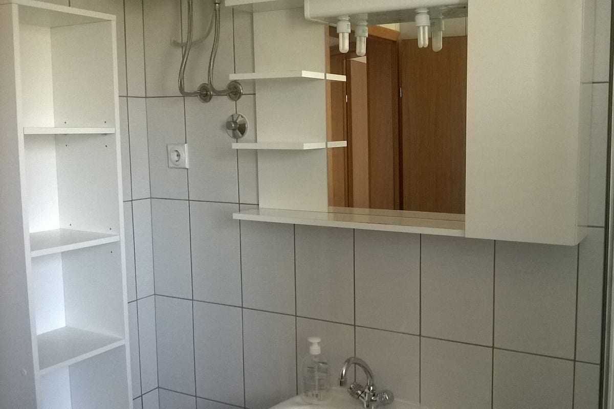 A bathroom with white tiled walls, a wooden vanity, a mirror, and a shower area.